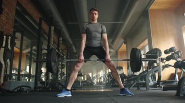 Spor salonunda halterle ağırlık kaldıran genç atletizm oyuncusuyla dolu. Sporcu halteri kaldırır, ayakları açıktır. Yüksek kalite 4k görüntü