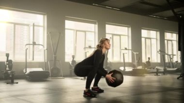 Sporcu bir kadın spor salonunda yerde tıbbi topla egzersiz yapıyor. Kadın topu göğsüne yakın tutuyor ve yere doğru iniyor. Yüksek kalite 4k görüntü