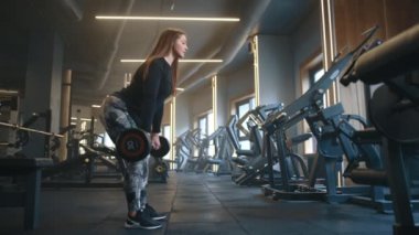 Formda genç bir kadının spor salonunda kaldırma hareketi yaparken çekilmiş profil fotoğrafı. Güçlü eğitim ekipmanlarıyla donatılmış. Kadın halteri kaldırıyor ve indiriyor. Düz görünüyor. Yüksek kalite 4k görüntü