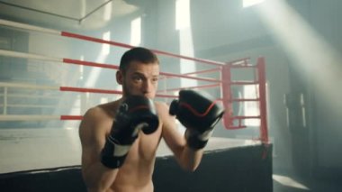 Boks eldivenli çıplak kaslı genç boksör ringin yanında dikilip gölge boksu yapıyor. Adam enerjik bir şekilde havaya yumruk atıyor. Yüksek kalite 4k görüntü 