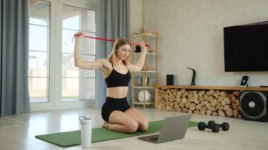 Çekici seksi bayan eğitmen çevrimiçi olarak evden eğitim veriyor. Kadın defter kullanıyor ve katılımcılara bakan direniş bandıyla egzersiz yapıyor. Yüksek kalite 4k görüntü