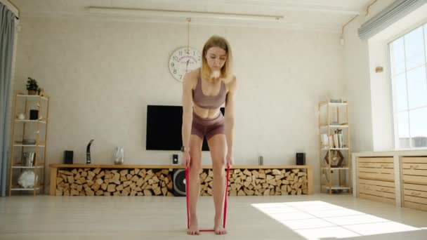 用电阻带练习 年轻的金发运动员在背直的情况下弯腰排成一排 女人在有现代室内装饰的公寓里锻炼 高质量的4K镜头 — 图库视频影像