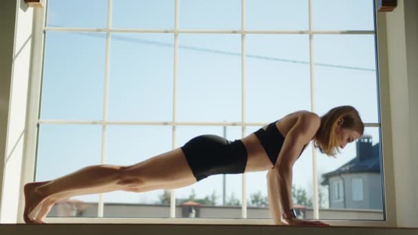 年轻的金发女运动员 光着细长的腿 在家里的大窗台上做俯卧撑 穿着黑色紧身衣的女人在窗边锻炼 高质量的4K镜头 — 图库视频影像