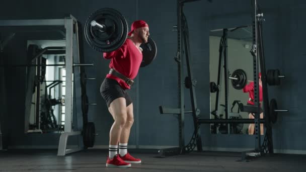 职业健美运动员蹲在体育馆里 带着杠铃 身穿红色衣服 动作缓慢 — 图库视频影像