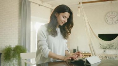 Oturma odasındaki masada tablete yazı yazan oldukça mutlu bir genç esmer. Hayalperest kadın gülüyor ve gülüyor, ev atmosferinde coşkuyla yazıyor. Yüksek kalite 4k görüntü