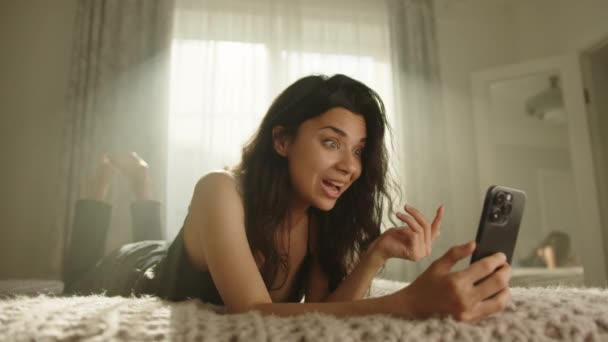 ビデオ通話による魅力的なブルネットは 日光に照らされた寝室の胃のベッドの上に横たわっています 若い女性が喜んで 幸せな驚いている 高品質4K映像 — ストック動画