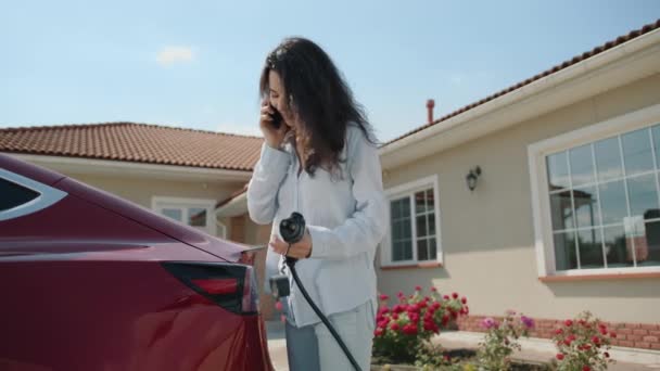 Roztomilá Kudrnatá Bruneta Nabíjí Červené Elektrické Auto Doma Blízkosti Jednopatrového — Stock video