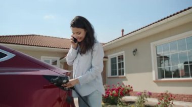 Genç esmer kadın, bir katlı modern evin yakınındaki avluda kırmızı elektrikli arabayı şarj ediyor ve cep telefonuyla konuşuyor. Kadın tel yerleştiriyor, gülüyor, saçlarını savuruyor. Yüksek kalite 4k görüntü