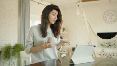 Çekici esmer, tablette oturup çay içerek video bağlantısıyla iletişim kuruyor. Genç bir kadın gülüyor, çok jestler yapıyor, kıvırcık saçlarını savuruyor. Yüksek kalite 4k görüntü