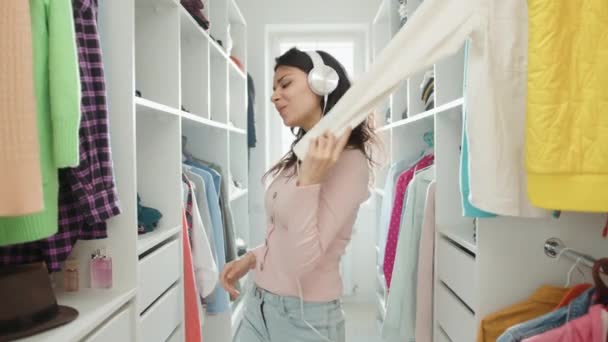 迷人的年轻黑发姑娘戴着耳机 在更衣室里唱歌跳舞 快乐的女人摸着衣服 戴上帽子 用胶辊当话筒 高质量的4K镜头 — 图库视频影像