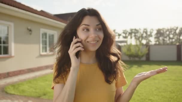 年轻快乐的黑发姑娘和一楼的房子在院子里散步 在手机上感情用事 绿色的草坪 后面是现代的私人住宅 高质量的4K镜头 — 图库视频影像