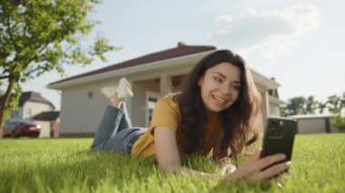 Gülümseyen esmer, yeşil çimlerin üzerinde uzanan telefondaki video bağlantısıyla iletişim kuruyor. Bir katlı özel bir evin bahçesinde yüzüstü yatan genç bir kadın. Yüksek kalite 4k görüntü