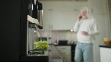 Modern mutfak tasarımının bulanık arka planında sade kahve makinesini kapat. Albino adam uzaktan geliyor ve kahve makinesine bir bardak koyuyor. Yüksek kalite 4k görüntü