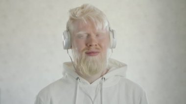 Beyaz kapüşonlu, beyaz kulaklıklı, beyaz arka planda en sevdiği şarkıyla dans eden genç sakallı albino bir adam. Olağanüstü bir adam. Yüksek kalite 4k görüntü