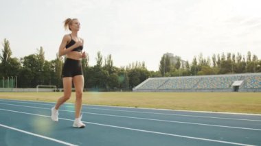 Dar şortlu güzel bir sarışın ve güneşli bir günde stadyumda koşuyor. Stadyum futbol sahası ve tribün koltuklarıyla donatılmıştır. Yüksek kalite 4k görüntü