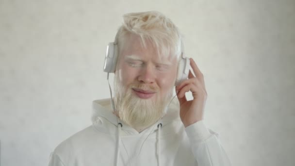 白いパーカーの髭を生やした若いアルビノ男性は 白い背景の上に白いヘッドフォンで音楽を聴く 異常な外観の幸せな男はリズムに移動します 高品質4K映像 — ストック動画