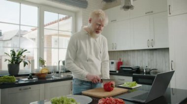 Yakışıklı, şık, sakallı albino adam evdeki büyük mutfak masasında kırmızı biber keserken bilgisayarında dostça bir video görüşmesi yapıyor. Yüksek kalite 4k görüntü