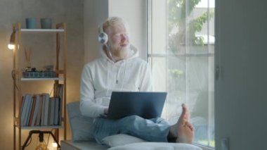 Beyaz kulaklıklı, dizüstü bilgisayarlı, geniş bacaklı, rahat bir odada pencere eşiğinde oturan albino bir adam. Adam pencereden dışarı bakıyor, gülümsüyor, rahatlıyor. Yüksek kalite 4k görüntü