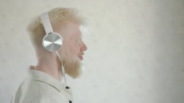 Beyaz kulaklıklı, gözleri kapalı genç, mutlu albino adam, beyaz arka planda müziğin ritmine doğru yavaşça ilerliyor. Profil fotoğrafı. Albinoların mutlu hayatı. Yüksek kalite 4k görüntü