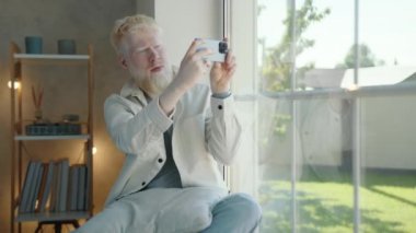 Albino adam akıllı telefondan dışarıyı gösteren video bağlantısıyla iletişim kuruyor. Genç adam, özel bir evin modern ve rahat bir odasında pencere eşiğinde oturuyor. Yüksek kalite 4k görüntü