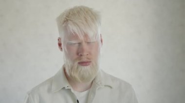 Kameraya bakarak gözlerini kısan genç albino adamın portresi. Göz albinizmi. Sakallı, mavi gözlü, beyaz tişörtlü, beyaz arka planda ceketi olan albino bir adam. Yüksek kalite 4k görüntü