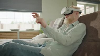 Genç heyecanlı albino adam VR kulaklık kullanıyor, dijital dünyada kendini eğlendiriyor, evdeki kanepede oturuyor. Albino adam dijital dünyada eylemlerini gerçekleştiriyor. Yüksek kalite 4k görüntü