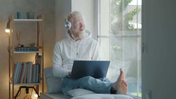 白化病患者 头戴白色耳机 膝上型笔记本电脑 双腿伸展 坐在舒适房间的窗台上 男人看着窗外 高质量的4K镜头 — 图库视频影像