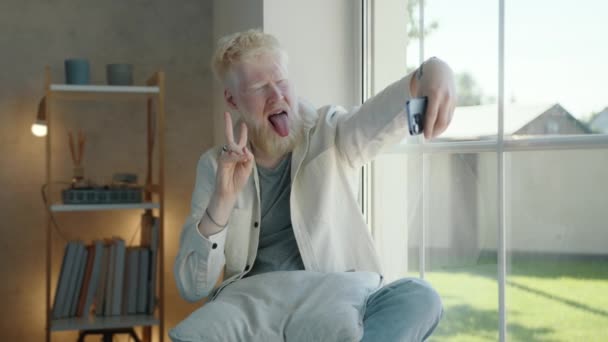 年轻的白化病患者做鬼脸 坐在窗边的智能手机上拍摄视频 积极的人表现出自己的舌头 发出和平的信号 高质量的4K镜头 — 图库视频影像