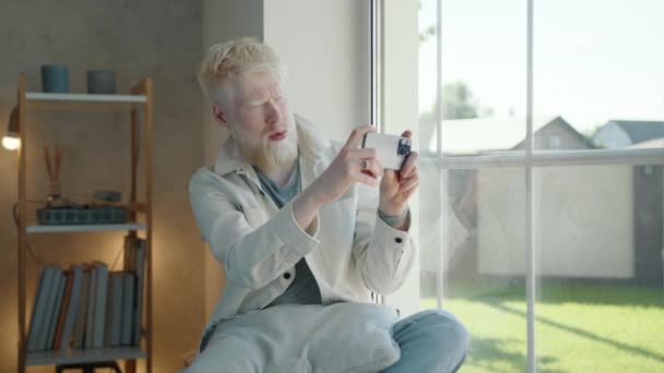 白胡子的白化病人坐在窗前拍户外风景的照片 年轻人打了几枪 对结果感到满意 无忧无虑的生活高质量的4K镜头 — 图库视频影像
