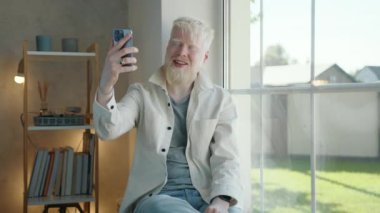 Heyecanlı stil sahibi albino adam evdeki akıllı telefondan video bağlantısıyla iletişim kuruyor. Adam el sallıyor, pencerenin kenarında oturuyor, iç ve dış manzarayı gösteriyor. Yüksek kalite 4k görüntü