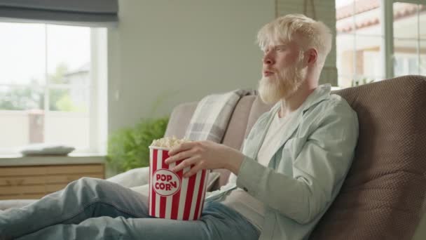 モダンなインテリアの大きな家のリビングルームで足を伸ばしてソファに座っているアルビノの男は ポップコーンを食べ テレビチャンネルを切り替えます いい家だ 高品質4K映像 — ストック動画