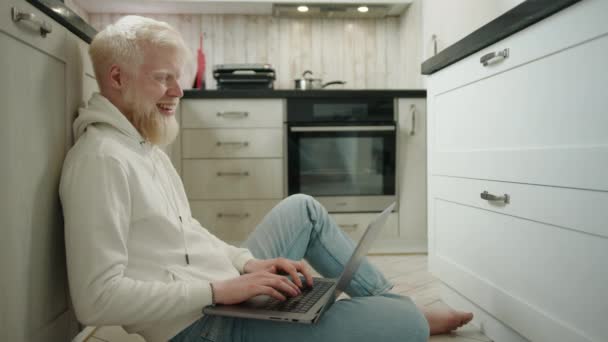 照片上年轻的白化病人 留着浓密的白胡子 穿着白色的帽衫和牛仔裤 坐在厨房地板上的笔记本电脑上打字 高质量的4K镜头 — 图库视频影像