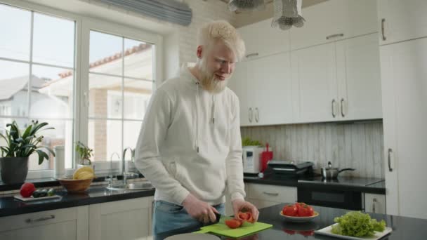 家のモダンなキッチンで緑のまな板にトマトを切るアルビノの男の笑顔 アルビニズムを持つ若いひげを生やしたきちんとした男は サラダを調理するのが好き クッカー 高品質4K映像 — ストック動画