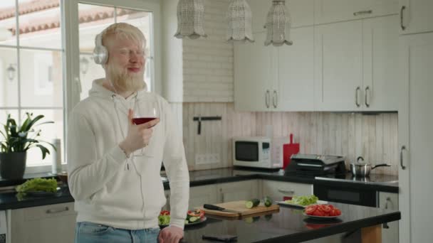 站在厨房里 戴着白色耳机 手里拿着一杯红酒 微笑着白化病患者 男人放松下来 听音乐 跟着节奏走 高质量的4K镜头 — 图库视频影像