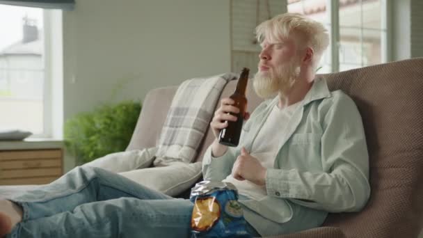 年轻胡子白化病人的闲暇时间 喝啤酒 吃薯片 坐在现代大房子客厅的沙发上 赢的姿势 足球赛 高质量的4K镜头 — 图库视频影像