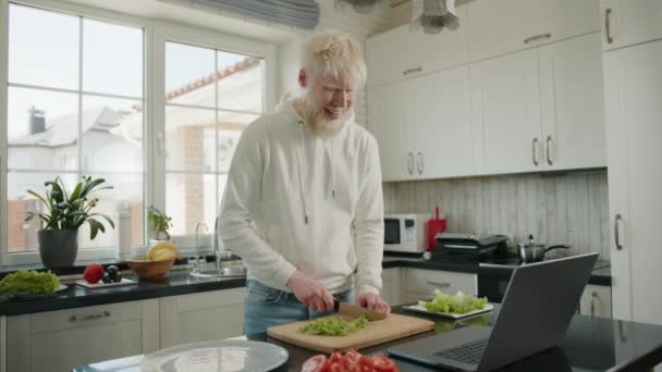 快乐的大胡子白化病人在现代厨房的切菜板上切生菜 通过笔记本电脑上的视频通话进行交流 白化病患者的生活方式高质量的4K镜头 — 图库视频影像