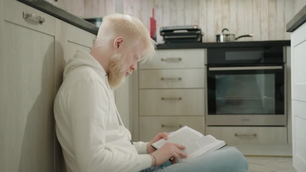 台所の床に孤独な座って本を読んでいるアルビノの男のプロフィールショット 台所で本を読むために皆から撤退するアルビニズムを持つ若い男 高品質4K映像 — ストック動画