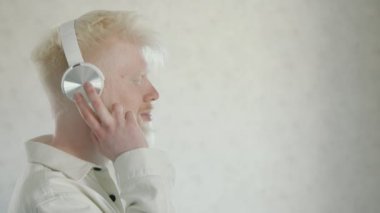 Beyaz kulaklıklı, gözleri kapalı, en sevdiği müziği dinleyen genç albino adamın profil fotoğrafı. Albinizmle sakallı adam şarkının ritmiyle dans eder. Yüksek kalite 4k görüntü