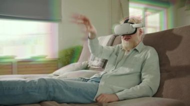 Heyecanlı albino adam, beyaz sakallı, VR kulaklıklı, ellerini havaya kaldıran, geniş odasındaki koltukta oturan, dijital dünyaya dalmış. VR etkisi içeriden geliyor. Yüksek kalite 4k görüntü