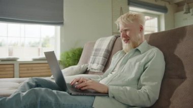 Albino adam evdeki kanepede dinleniyor, bilgisayardaki video bağlantısıyla iletişim kuruyor. Albino adam el sallıyor, video aracılığıyla hoş sohbetler yapıyor, kanepede oturuyor. Yüksek kalite 4k görüntü
