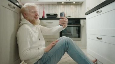 Mutlu albino adam mutfakta yerde oturmuş akıllı telefonuyla video görüşmesi yapıyor. Yakışıklı albino adam merhaba diyor ve cep telefonundan sohbetin tadını çıkarıyor. Yüksek kalite 4k görüntü