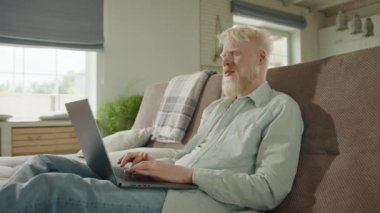 Dizüstü bilgisayarında yazı yazan, geniş oturma odasında bacakları esnemiş, ciddi bir albino adam. İnternet işi. Sosyal zorluklar. Yüksek kalite 4k görüntü