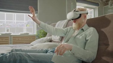 Sanal gerçeklik gözlüklü albino adam oturma odasında kanepede oturuyor. VR etkisi içeriden geliyor. Albino dijital dünyada yaşarken ellerini önünde gezdiriyor. Yüksek kalite 4k görüntü