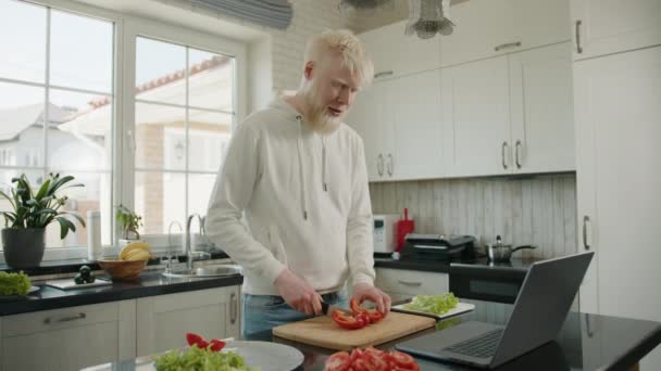 ラップトップでビデオ会議をしている間に台所で唐辛子を切る若いアルビノの男 自宅で料理をしながら アルビニズムを持つ男は ビデオリンクを介して通信します 高品質4K映像 — ストック動画