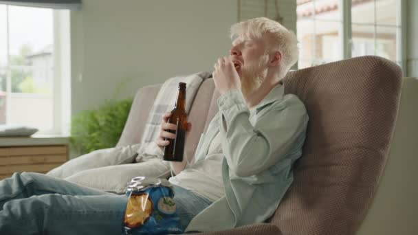 알비노 남자는 맥주를 마시고 집에서 휴식을 취하고 다리가 늘어진 소파에 — 비디오