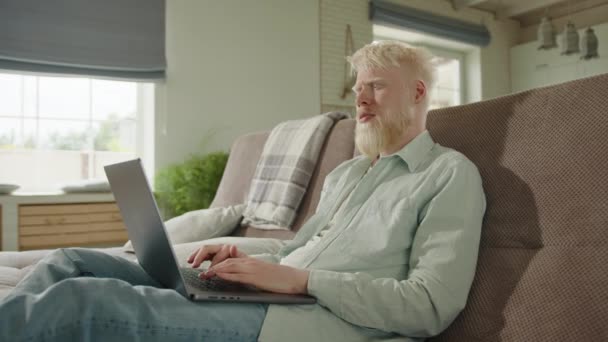 严重的白化病患者 白色浓密的胡子在笔记本电脑上打字 坐在沙发上 伸展的腿在宽敞的客厅里 在线工作 社会挑战 高质量的4K镜头 — 图库视频影像