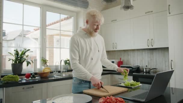 ラップトップのビデオリンクを介して通信するアルビノの男 現代のキッチンで料理 アルビニズムの波がこんにちは男とまな板の上にレタスの切断を開始します 高品質4K映像 — ストック動画
