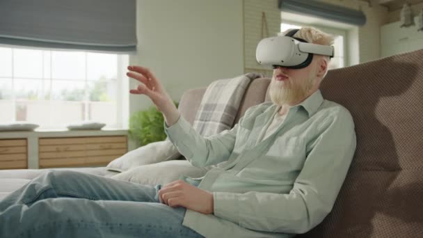 白化病患者戴着白色虚拟现实眼镜坐在客厅的沙发上 白化病人的手在他面前移动 沉浸在数字世界里 高质量的4K镜头 — 图库视频影像