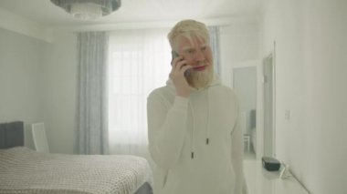 Kalın beyaz sakallı, cep telefonuyla konuşan, beyaz iç tasarımı olan ferah bir evde yürüyen mutlu genç albino adam. Albinoların ev hayatı. Yüksek kalite 4k görüntü