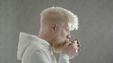 Orta boy, profil çekiminde albino adam sıcak içeceği tadıyor. Beyaz kapüşonlu olağandışı bir adam sıcak çay içer, zevkle gözlerini kapatır. Beyaz arka plan. Yüksek kalite 4k görüntü